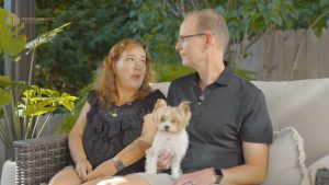 Couple on outdoor sofa under Pergomatic pergola; man in black polo holds small white-brown dog while chatting with partner.