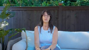 Pergomatic homeowner in light blue top and pink glasses shares testimonial on patio sofa against dark wood fence.