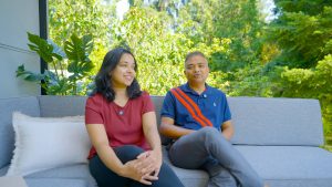 Smiling couple on grey outdoor sofa discusses Pergomatic pergola; red blouse and navy stripe polo pop against green backdrop.