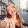 Closeup selfie-portrait pretty blonde girl on street in city. She has vinous lips, smiling to camera
